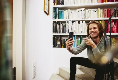 Smiling man sitting on stairs, making a selfie. bookshelf in background