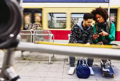 male and female looking at phone while sitting on a bench on a train platform.
