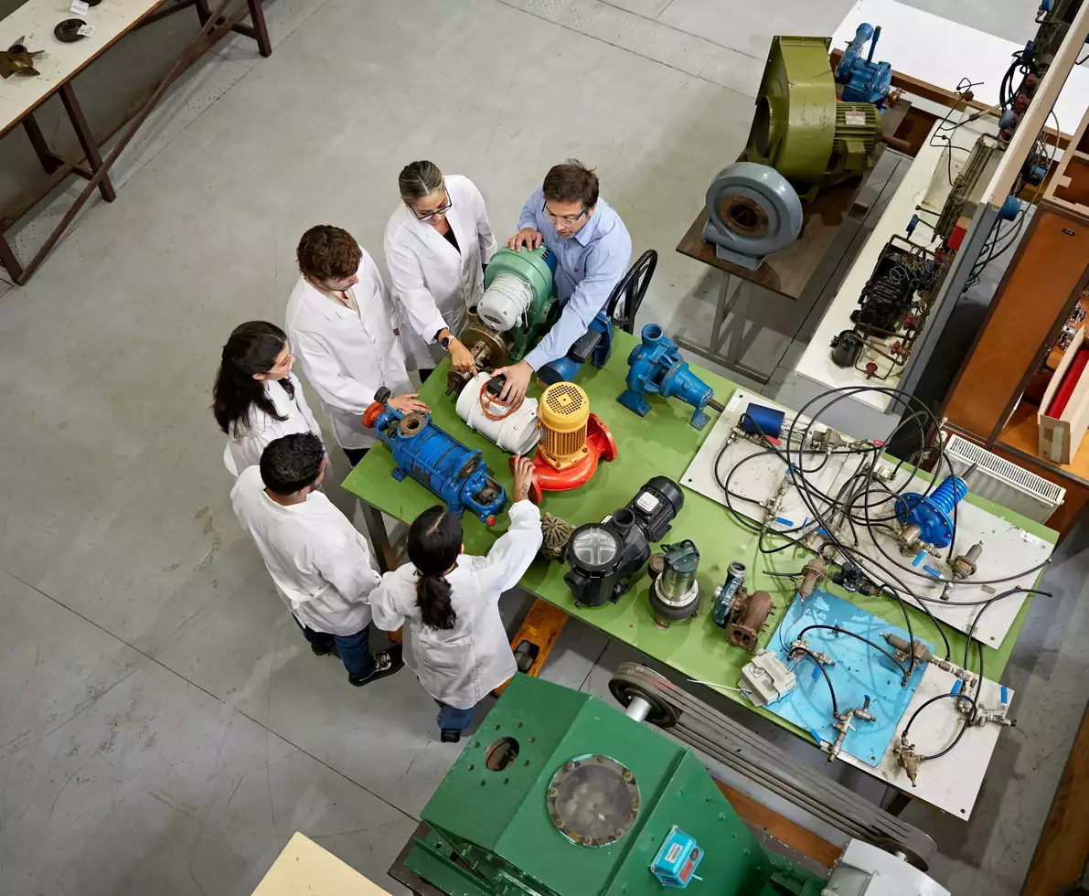 High angle view of mature man and group of students in lab coats gathered around a table and learning about tools and materials.